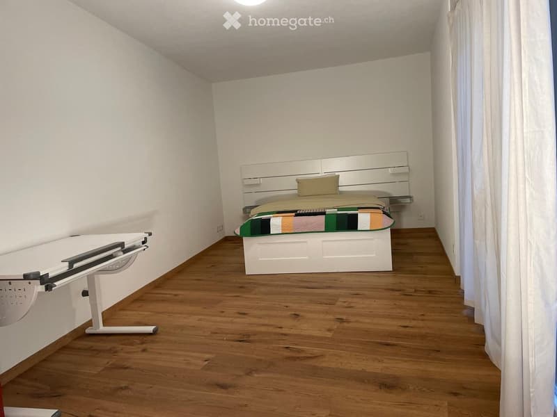 Modern bedroom with wooden flooring, white walls, a bed with a colorful quilt, and a white desk.