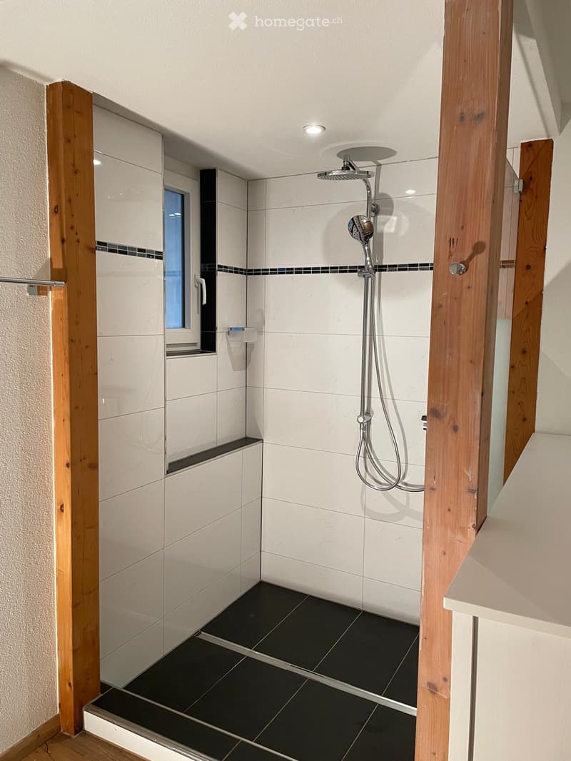 Modern shower area with white tiles, black accents, and a glass enclosure. Features a handheld showerhead, a built-in shelf, and a small window for natural light.