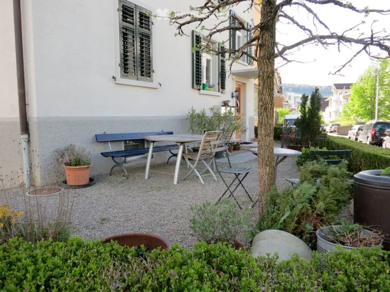 A small, well-maintained garden area with a seating set including a table, chairs, and a bench, surrounded by potted plants, shrubs, and a tree. The area is paved with gravel and features a white building with shutters in the background.