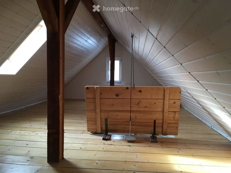 An attic space with wooden floors and exposed wooden beams, featuring skylights and a large wooden chest suspended from the ceiling.