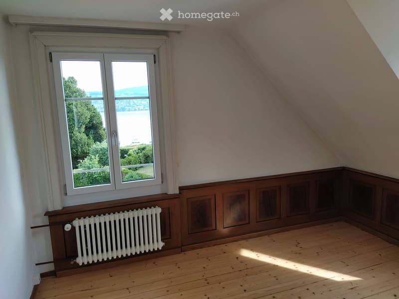 Empty attic room with wooden floor, slanted ceiling, large window offering a scenic view, and a white radiator below the window.