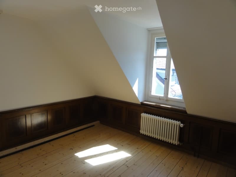 Empty attic room with wooden floor, slanted ceilings, white walls, and a single window with radiator below.