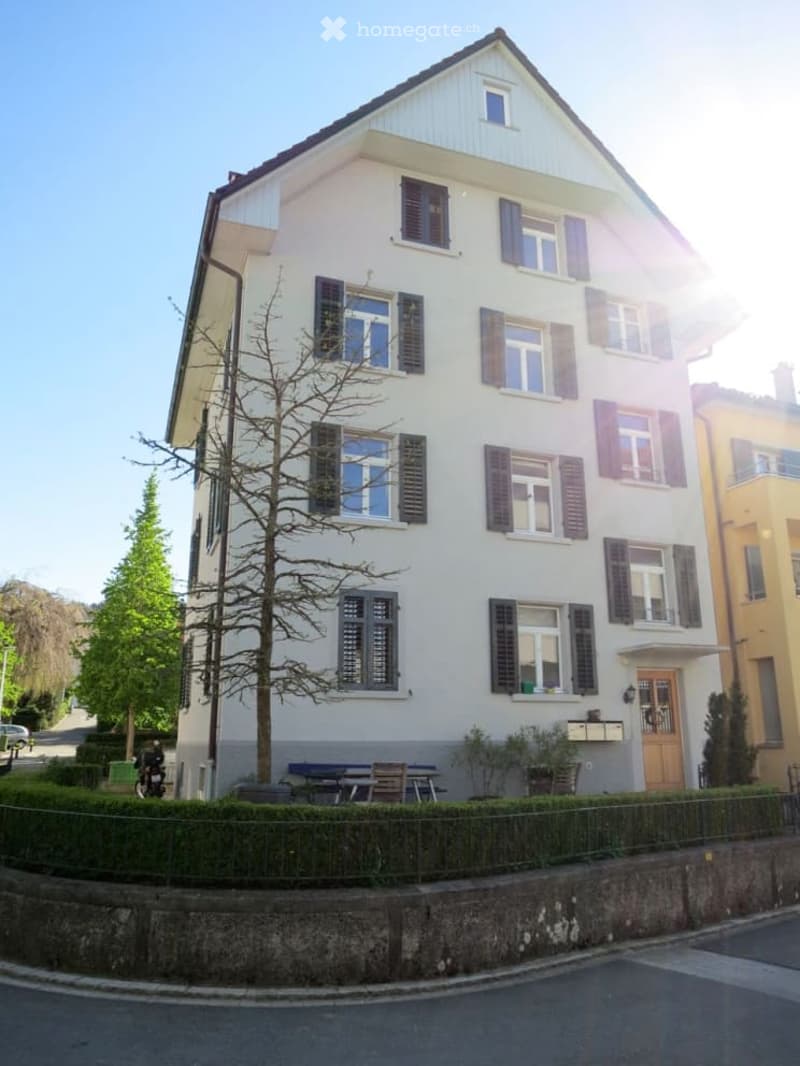 Two-story house with white exterior, multiple windows with shutters, and a small garden area with a bench and table.