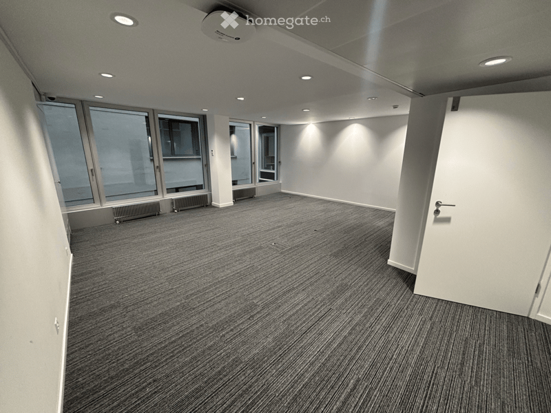 Empty room with grey carpeted floor, white walls, large windows, and recessed lighting.