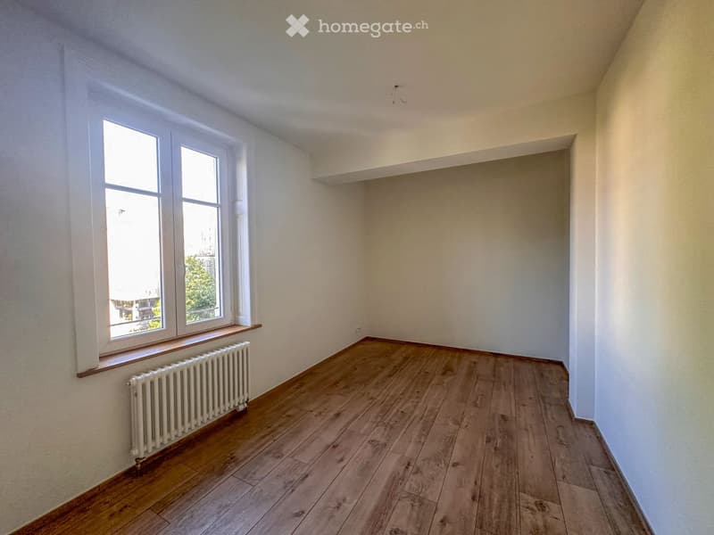 Empty room with wooden flooring, white walls, large windows, radiator, and a small shelf.