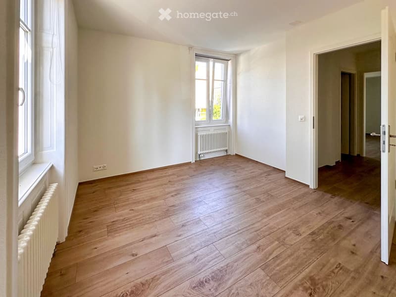 Empty room with wooden flooring, white walls, and large windows. Radiators are visible along the walls, and there is an open doorway leading to another room.