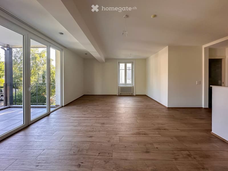 Empty room with wooden flooring, white walls, large sliding glass doors leading to a balcony, and a small window with a radiator below.