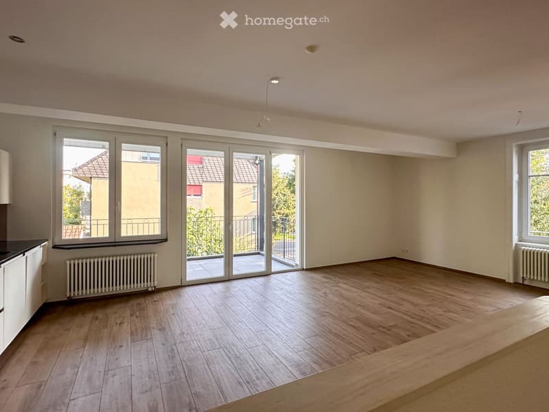 Empty room with wooden flooring, large windows, sliding glass doors leading to a balcony, and radiators on the walls.