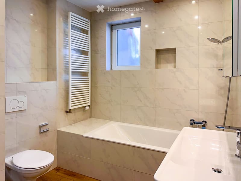 Modern bathroom with marble tiles, a bathtub, a toilet, a sink, a radiator, and a mirror. The room features a small window and a sleek faucet design.