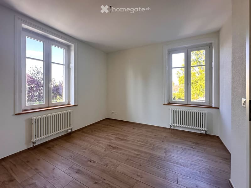 Empty room with wooden flooring, white walls, and large windows with radiators beneath them.
