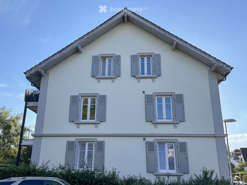 Two-story house with white exterior, grey shutters, multiple windows, and a small balcony on the left side.
