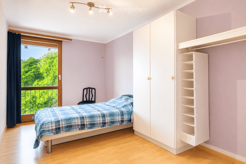 Cozy bedroom with a single bed, a chair, a wardrobe, and a shelf. The room features wooden flooring, light purple walls, and a large window with a view of greenery.