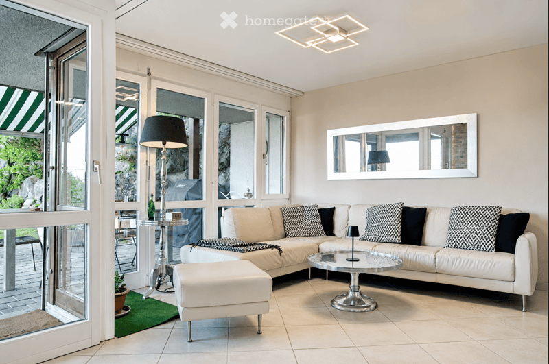 Modern living room with a white sectional sofa, coffee table, ottoman, floor lamp, side table, and a large mirror on the wall. The room features large windows with a view of a garden and has a sleek, contemporary design.