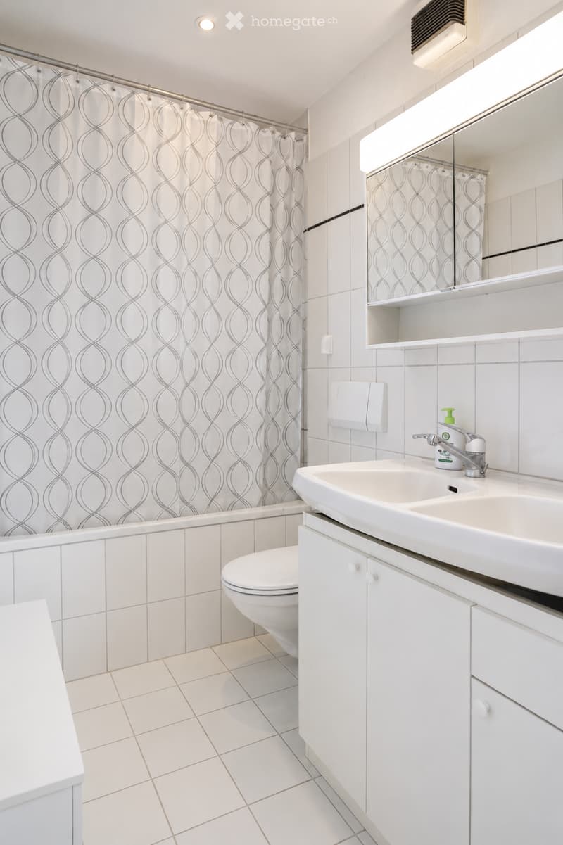 Modern bathroom with white tiles, a white sink with a chrome faucet, a toilet, a mirrored cabinet, and a shower with a decorative curtain.