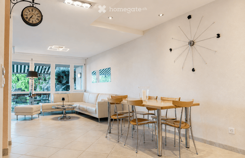 Bright living and dining area with white walls, tiled floor, large windows, a white sectional sofa, a glass coffee table, a wooden dining table with chairs, and decorative lamps.
