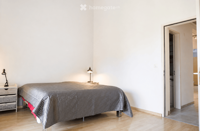 Modern bedroom with a large bed covered in a grey duvet, a nightstand with a lamp, and a drawer unit. The room features light wooden flooring and white walls.