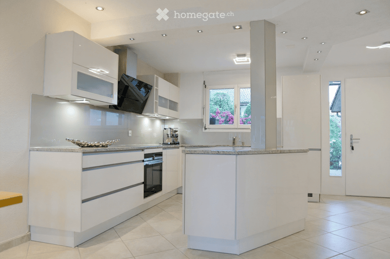 Modern kitchen with white cabinets, granite countertops, built-in oven, microwave, and coffee maker. Features a central island with a sink and faucet, and a window with a view of greenery.