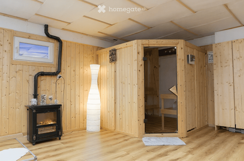 Cozy room with wooden walls and floor, featuring a black fireplace, a tall white vase, a small table with glasses, and an open wooden sauna with benches inside.