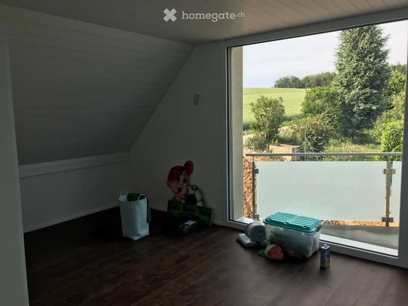 Bright, wooden-floored room with large windows offering a scenic view of greenery. Contains a basket, storage bin, toy, books, and a cup.
