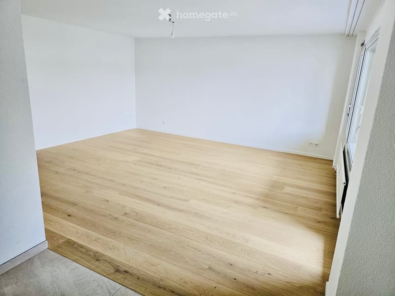 Empty room with light wooden flooring and white walls, featuring a window and a door.