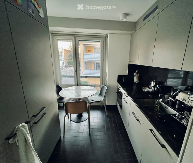 Modern kitchen with white cabinets, black countertops, and a small dining area with a round table and chairs. Appliances include an oven, fridge, and coffee machine.