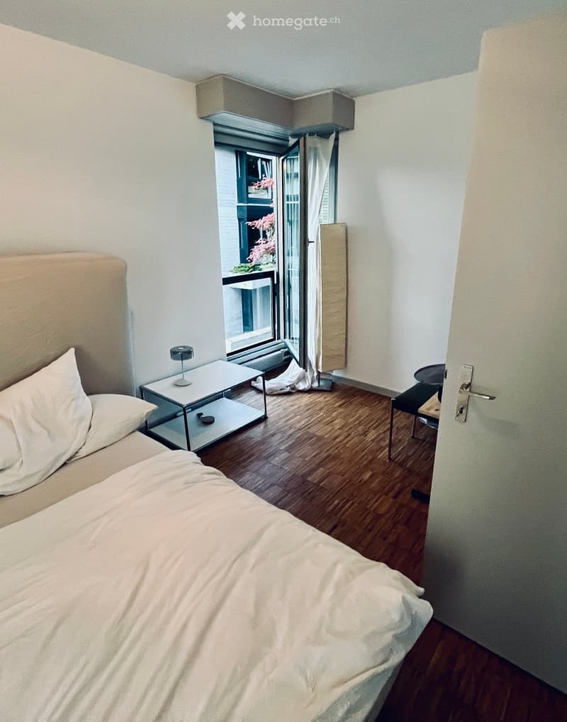 Cozy bedroom with a large bed, nightstand, small table, and chair. Features wooden flooring, white walls, and a large window with a view of a balcony.