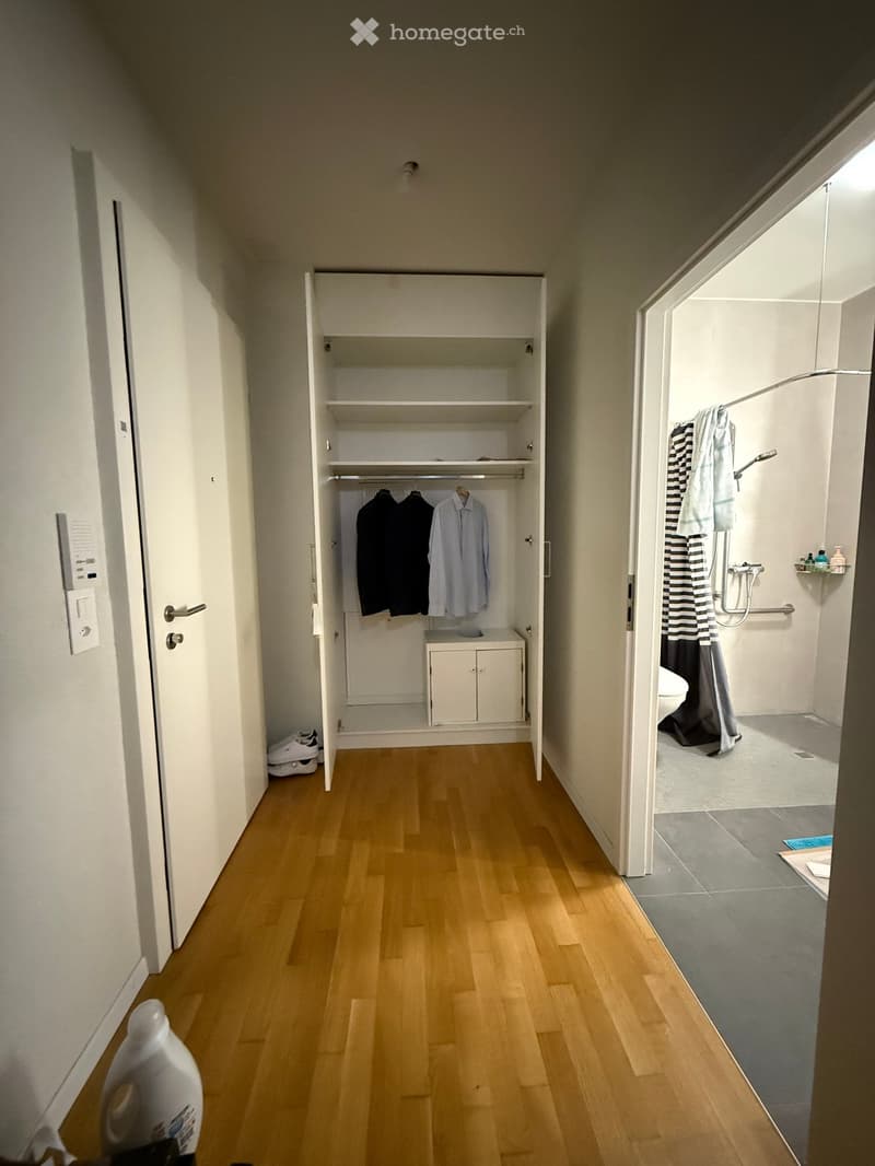A narrow hallway with wooden flooring, leading to a closet and a bathroom. The closet contains hanging clothes and shelves, while the bathroom has a shower, towel, and toiletries.