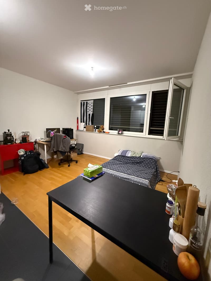 A furnished room with a bed, a table, a chair, a computer, a backpack, a cupboard, a fridge, and kitchen utensils.