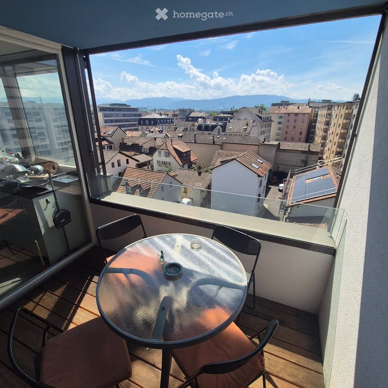 Balcony with a round glass table and two chairs, overlooking a cityscape with rooftops and distant mountains.