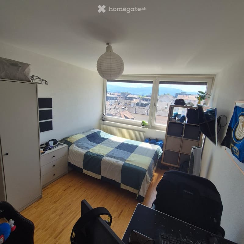 Cozy bedroom with a double bed, nightstand, desk, and chair. Large windows offer a scenic city view. Includes a wardrobe, fan, and various personal items.