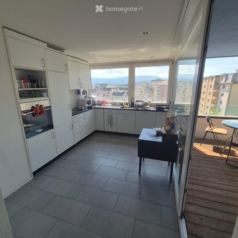 Modern kitchen with white cabinets, tiled floor, and large windows offering a scenic view of the city and mountains. Includes an oven, microwave, fridge, sink, and a small table with chairs on the adjacent balcony.