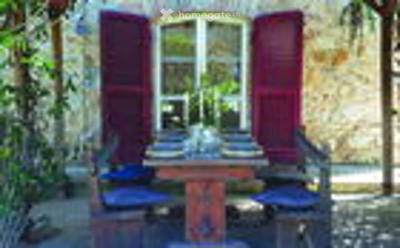 A small outdoor dining setup with a wooden table and chairs, surrounded by greenery and a wooden fence.