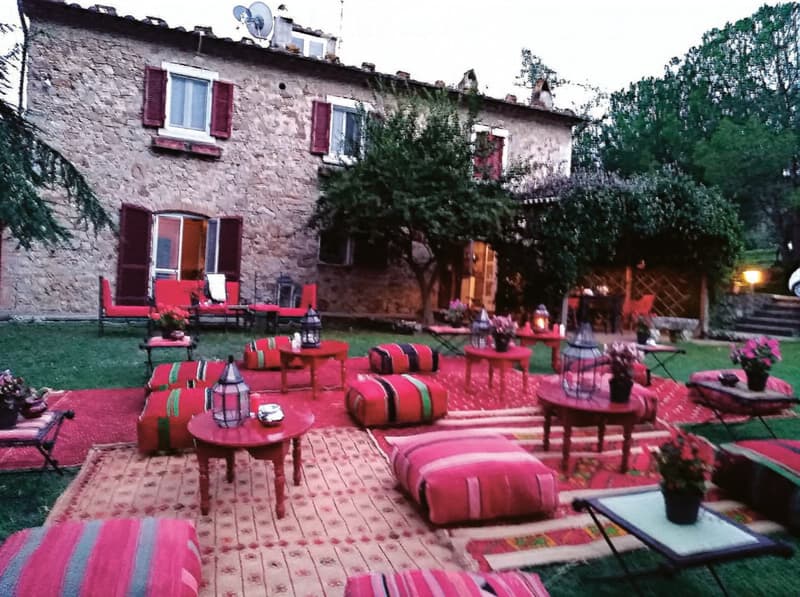 Outdoor seating area with red and pink cushions, small tables, lanterns, and flower pots, set on patterned rugs in front of a stone building.