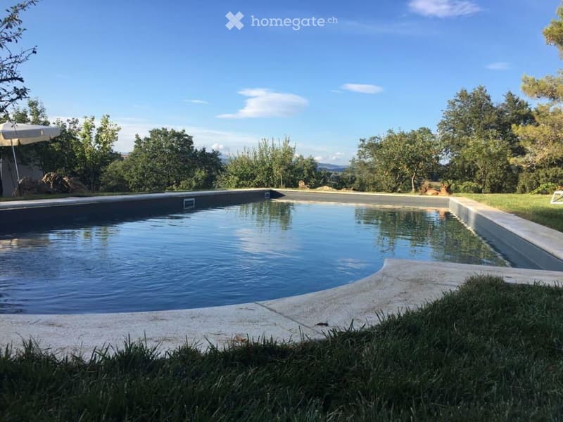 A rectangular swimming pool surrounded by a concrete deck, with grassy areas and trees in the background under a clear blue sky.