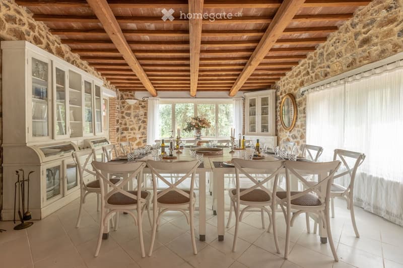 Dining room with exposed wooden beams, stone walls, large windows with white curtains, a round wooden dining table surrounded by white chairs, cabinets, and a decorative mirror on the wall.