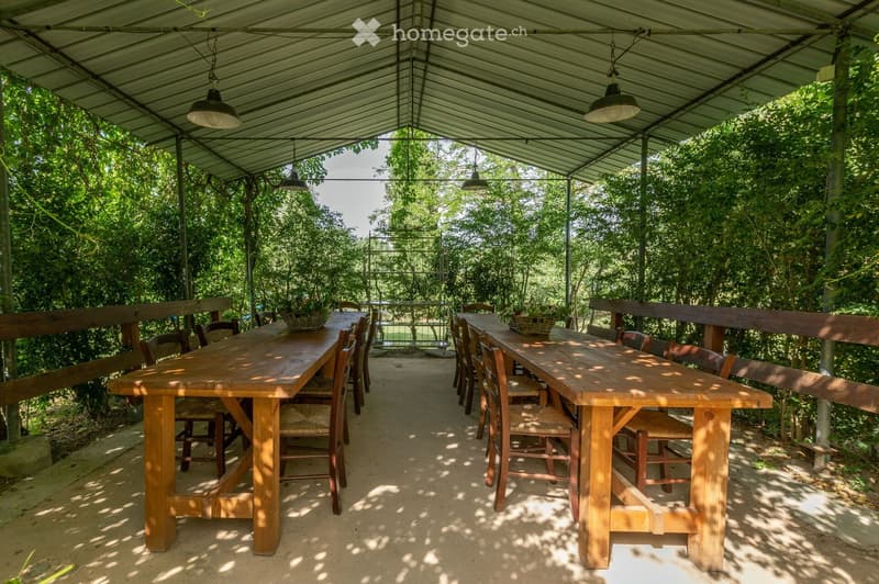 An outdoor dining area with two long wooden tables and multiple chairs, surrounded by lush greenery and trees, with hanging pendant lights under a metal roof structure.