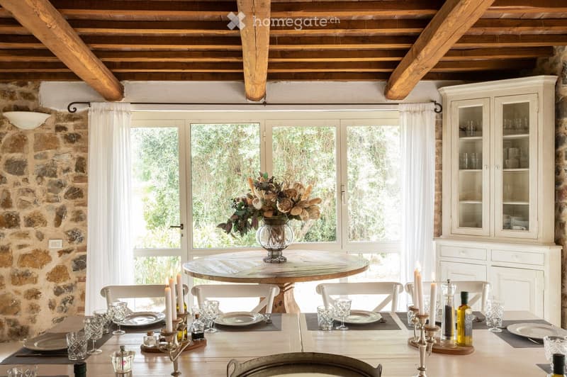 Dining room with a round wooden table, white chairs, a vase with flowers, and a cabinet with glass doors.