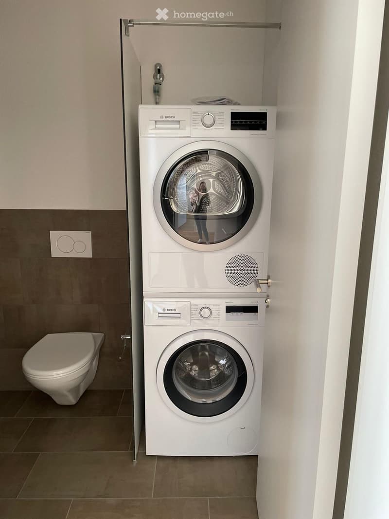 Modern bathroom with a white Bosch washing machine and dryer unit, a toilet, and tiled walls and floor.