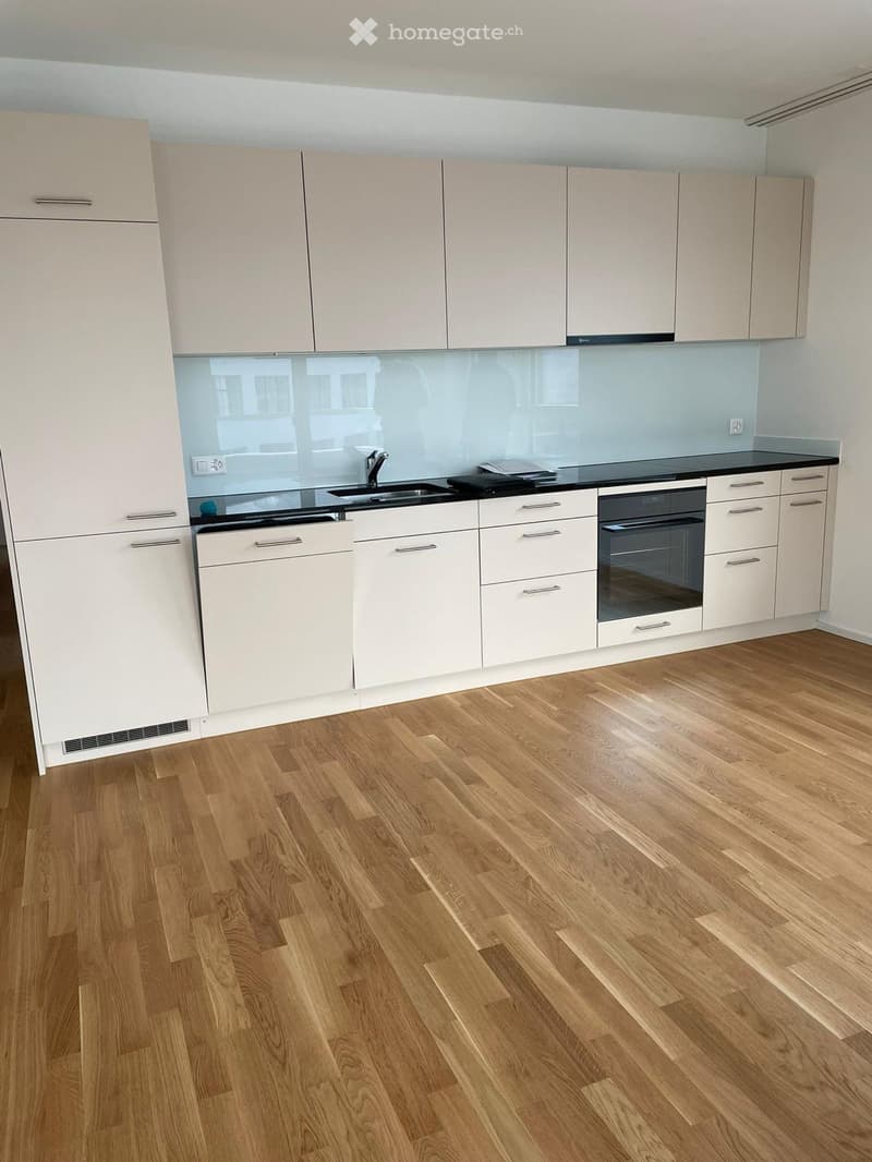 Modern kitchen with light-colored cabinets, wooden flooring, and a black countertop. Features a sink, faucet, and built-in oven.
