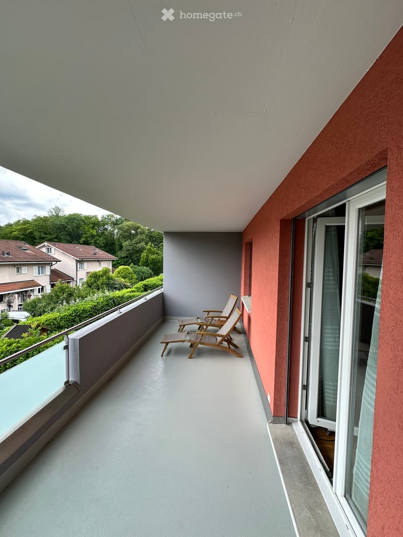 Balcony with two wooden chairs and a small table, overlooking a green landscape with houses and trees.