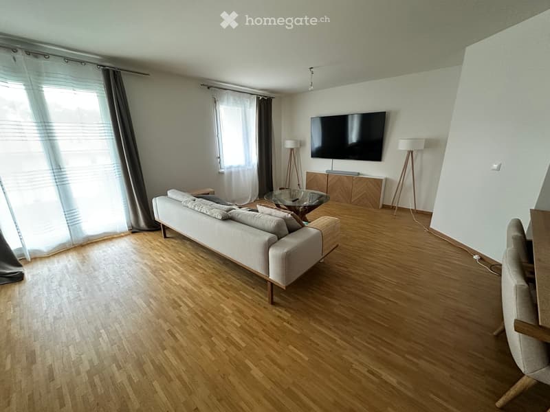 Modern living room with light wooden flooring, a grey sofa, a glass coffee table, a television mounted on the wall, and two floor lamps.