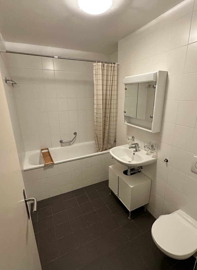 Modern bathroom with white tiles, bathtub, sink, toilet, mirror, and a small wooden tray on the edge of the bathtub.