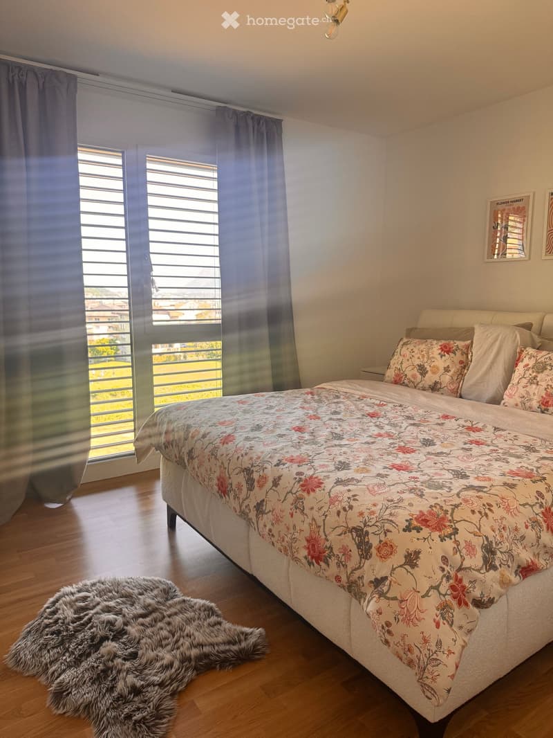 Bedroom with a large bed, floral bedding, and a fur rug on the floor. Large windows with blinds and curtains provide natural light.