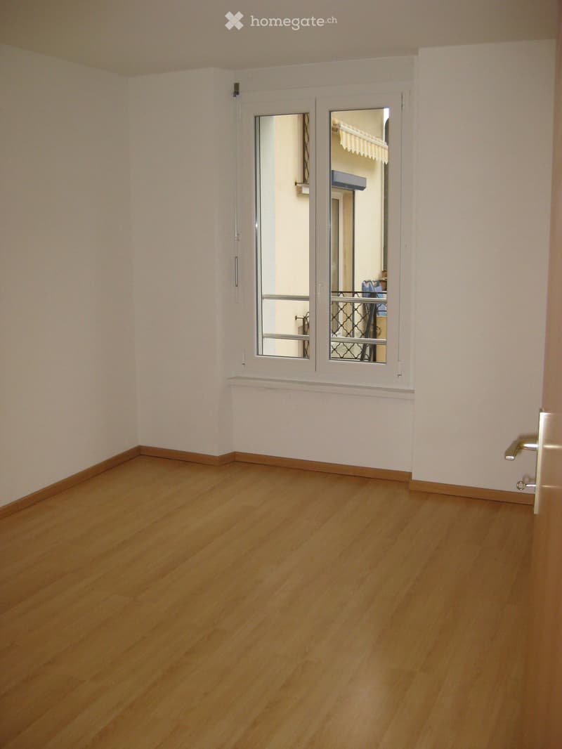 Empty room with light wooden flooring and white walls, featuring a large window with a view into an adjacent room.