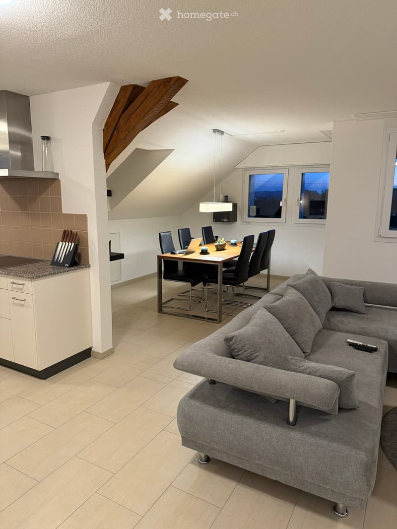 Modern, open-concept living and dining area with a grey sectional sofa, a dining table with six chairs, a laptop, and a small kitchen area featuring a countertop, cabinets, and a knife block. The space has large windows and a slanted ceiling with exposed