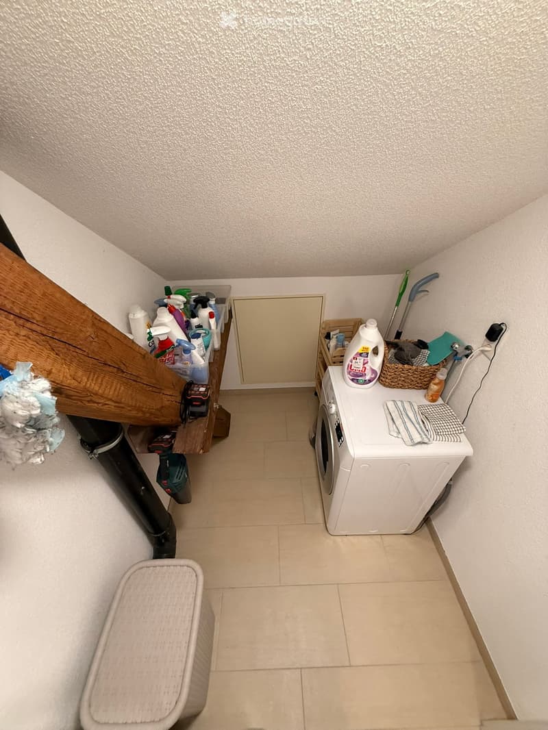 Small laundry area with a washing machine, laundry basket, cleaning supplies, and a stool.