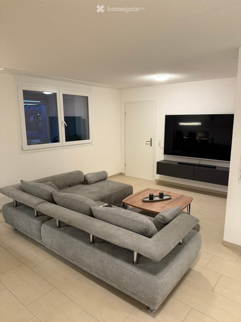 Modern living room with a grey sectional sofa, wooden coffee table, flat-screen TV on a stand, and large windows.