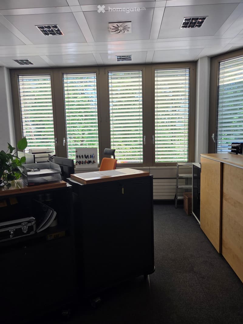 Office space with desks, chairs, a plant, and a printer, featuring large windows with blinds and a ceiling with ventilation grilles.