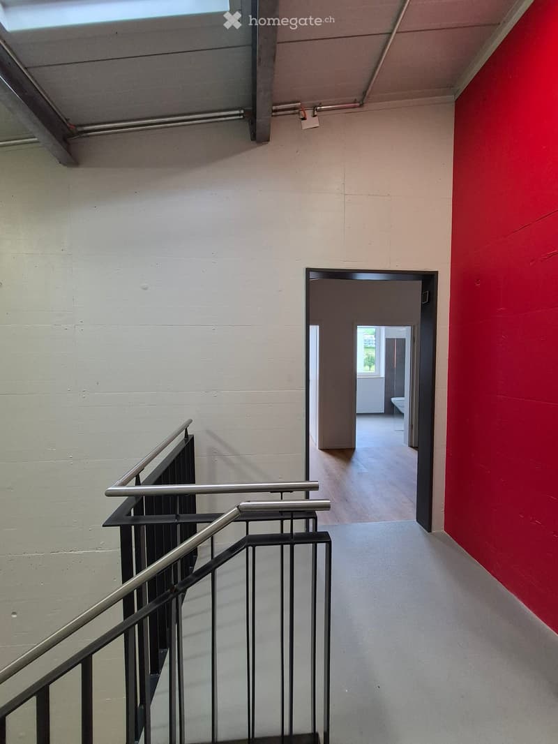 Modern hallway with a staircase, metal railings, and a red accent wall. A doorway leads to a room with a visible toilet.