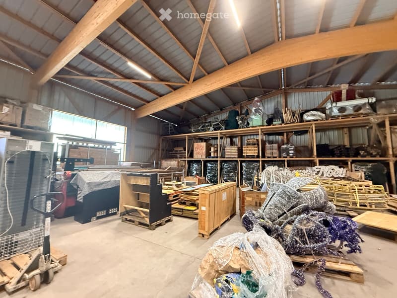 Large, open warehouse space with exposed wooden beams and metal roofing. Shelves hold various items, including boxes, tools, and materials. A forklift is visible on the left side.